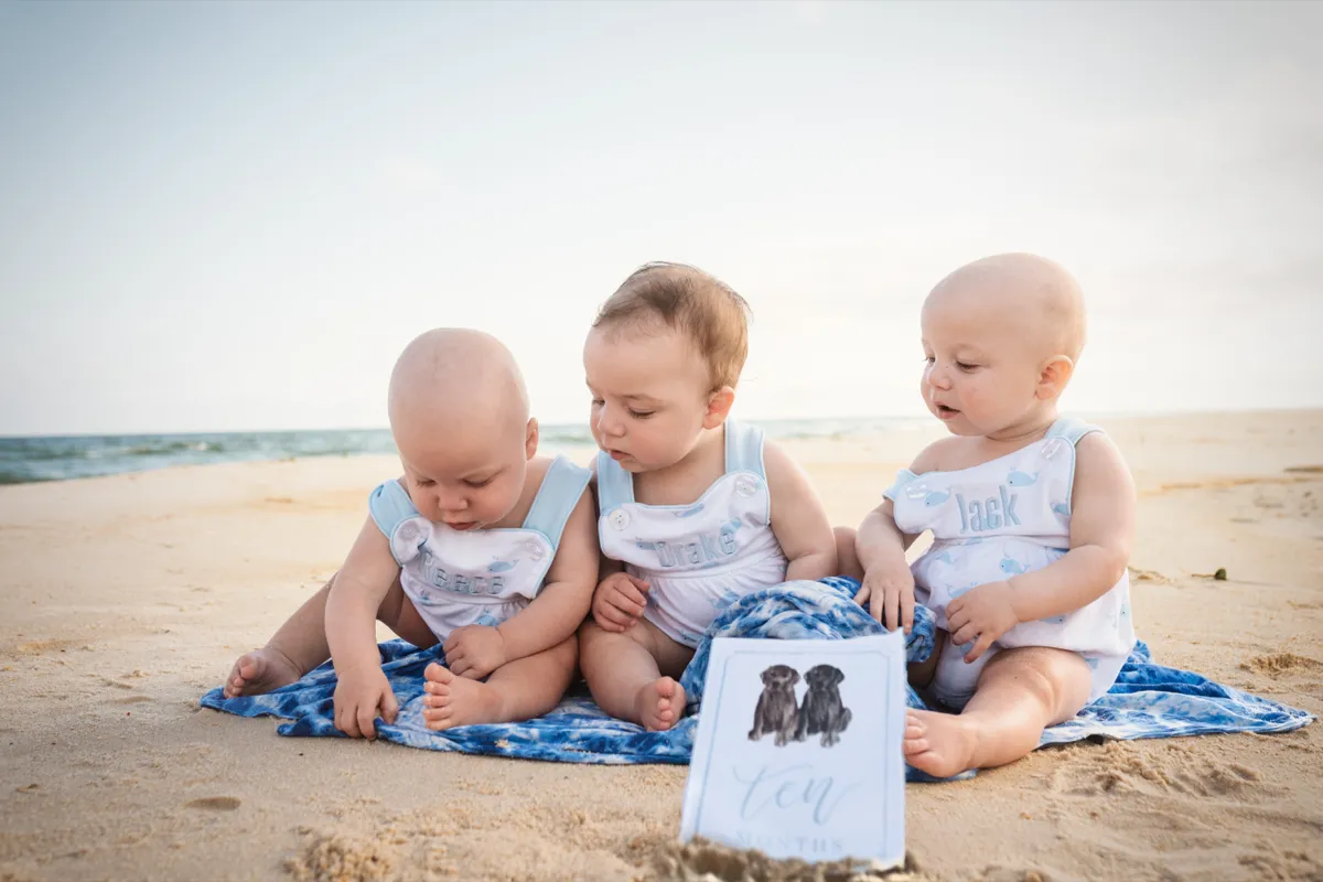 Three babies in matching outfits sitting together on a blanket at the beach