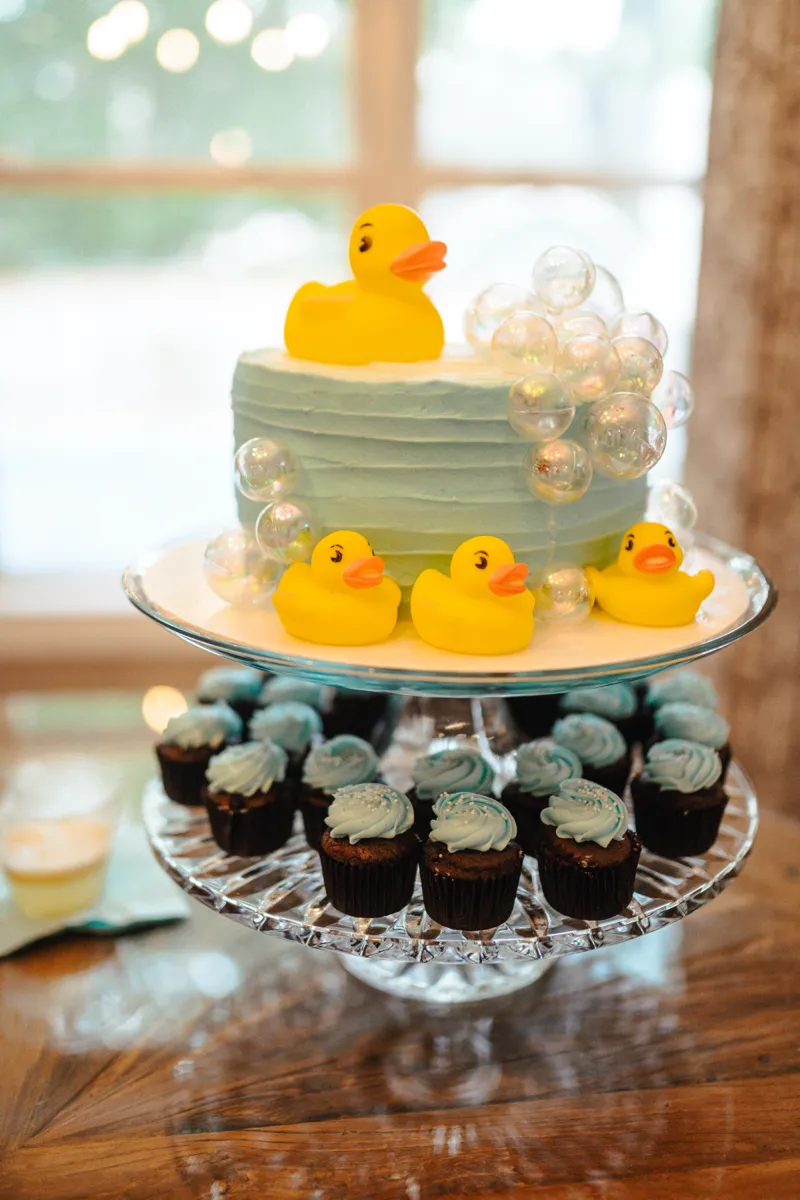 A pale blue baby shower cake topped with a rubber duck, above a tier of frosted cupcakes