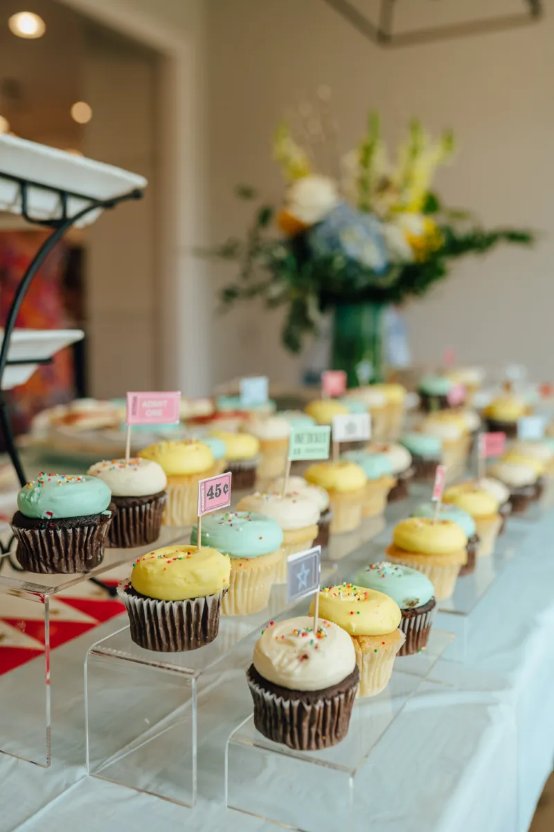 A long table of pastel cupcakes with hand-lettered labels and a flower arrangement behind