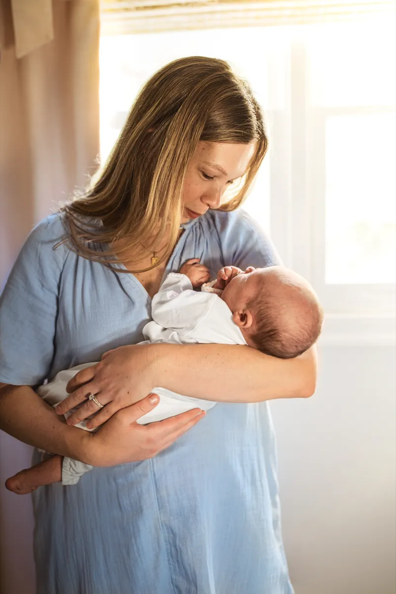 A mother in a blue dress holding her sleeping newborn beside a sunlit window