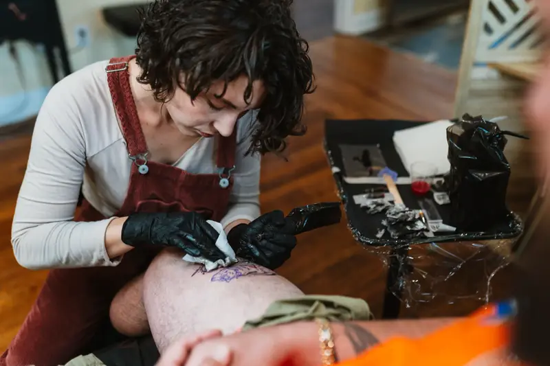 A tattoo artist in a red apron leaning in to work on a client's leg