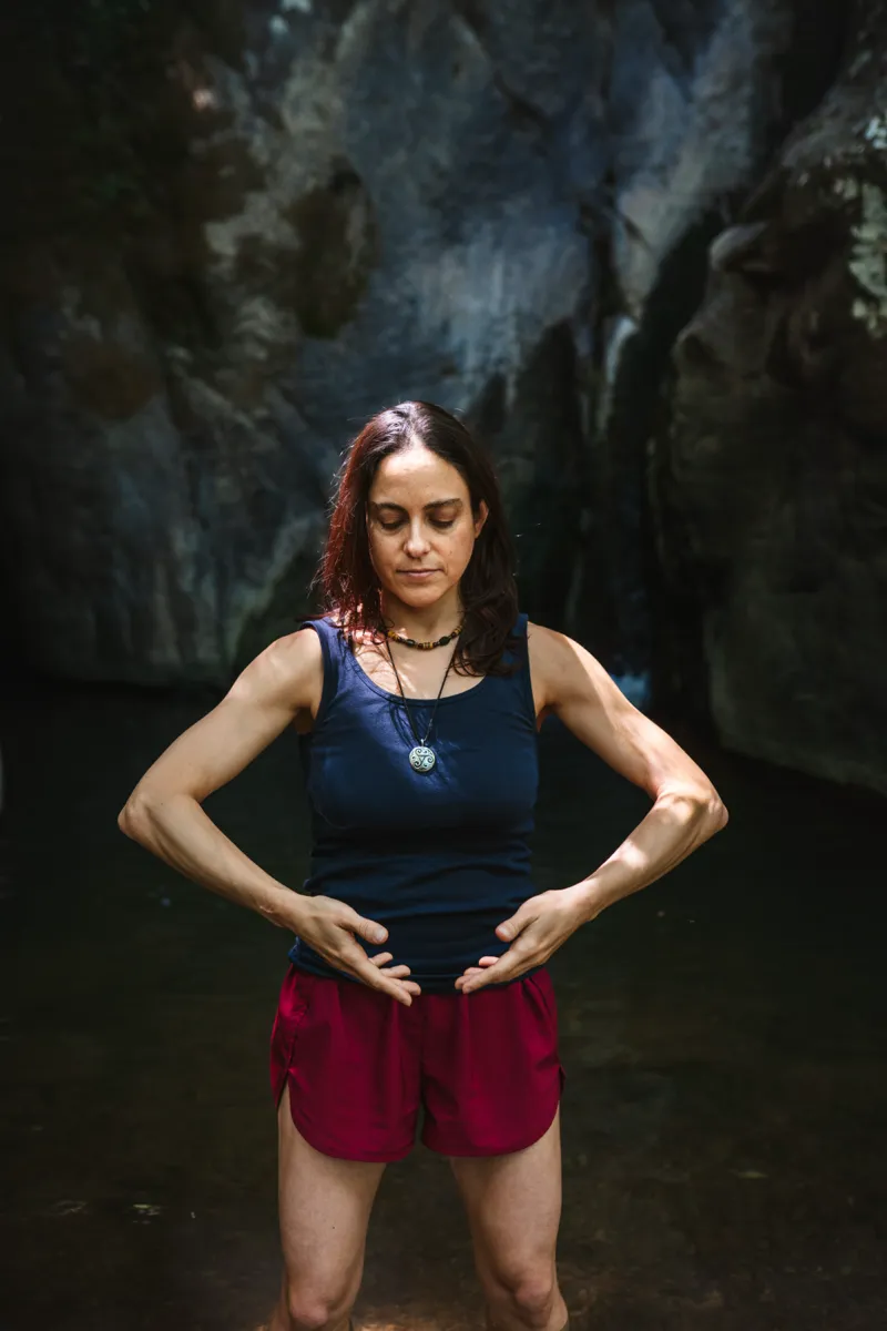 A woman standing in shallow water beneath a rock face, hands at her waist, eyes closed