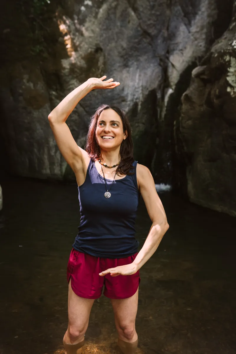 A woman practising a raised-arm movement in shallow water beneath a rock face