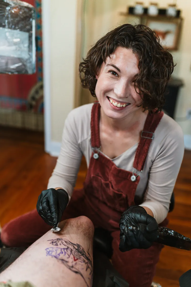 A tattoo artist smiling toward the camera, mid-session, gloved hands at work