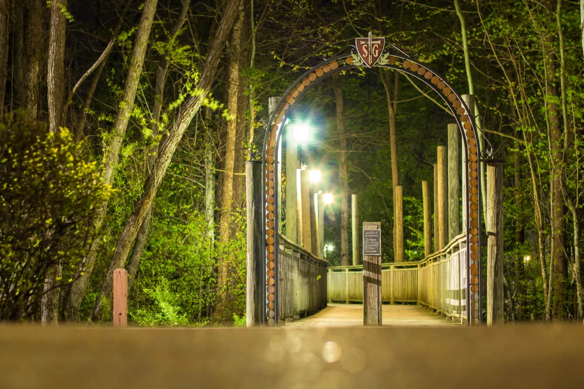 A lit wrought-iron arch marking the entrance to a wooden boardwalk through the woods at night