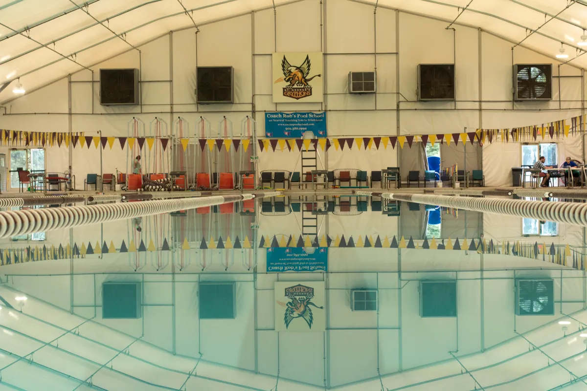 A competition pool under a white tented roof, lane lines and pennant flags mirrored in still water