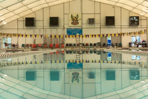 A competition pool under a white tented roof, lane lines and pennant flags mirrored in still water