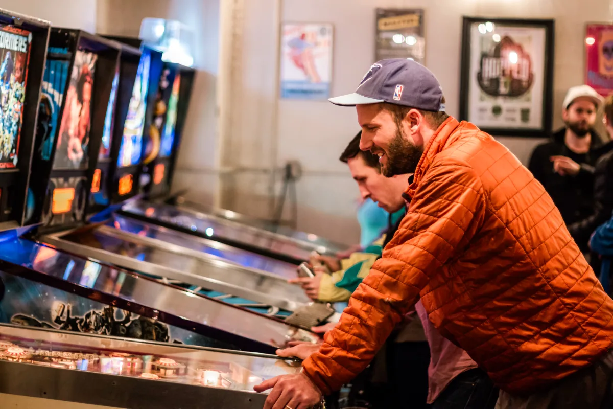 A player grins mid-game, hands on the flipper buttons, lit machines beside him.