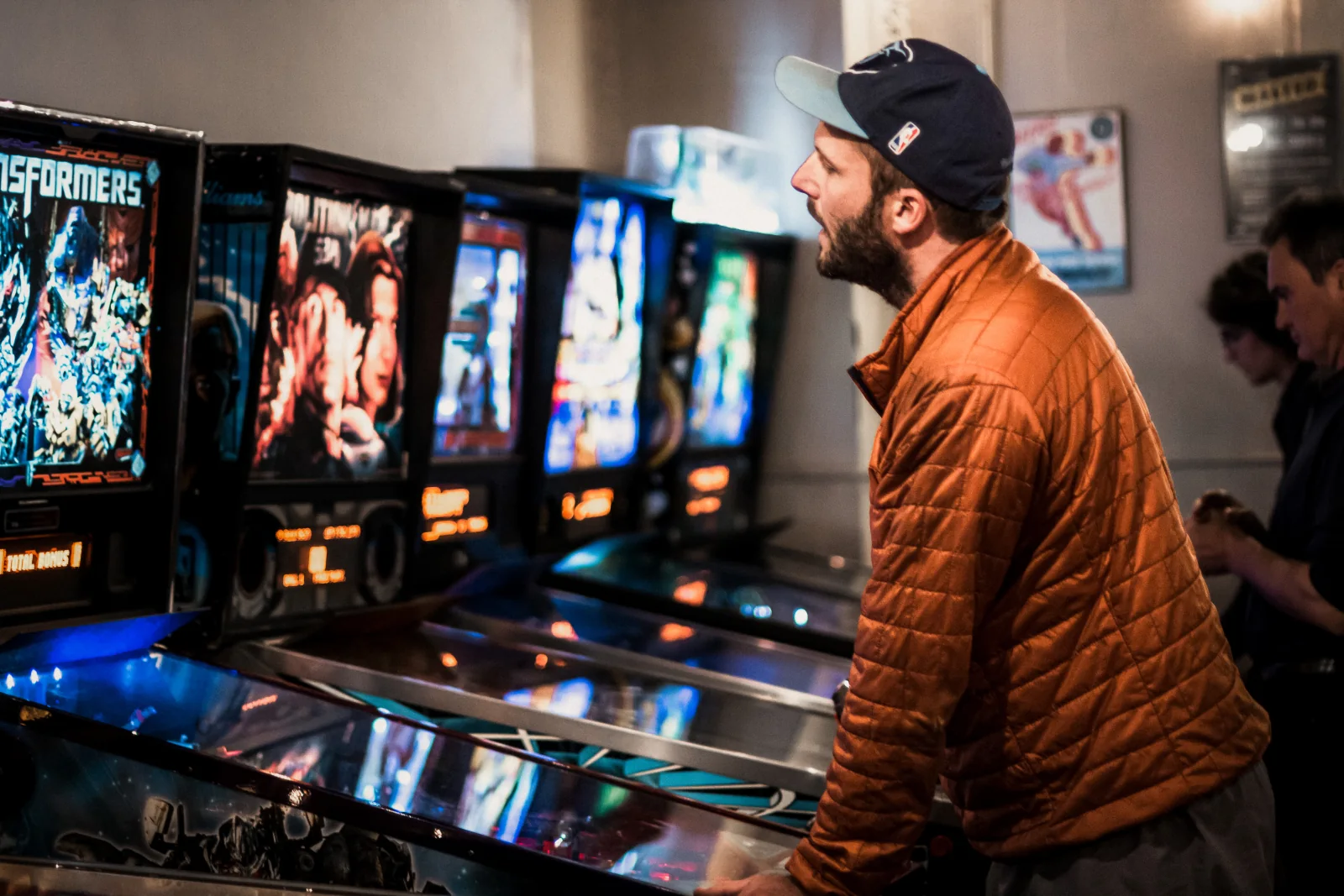 A player in an orange jacket leans over a machine at the end of a lit row of pinball backglasses.