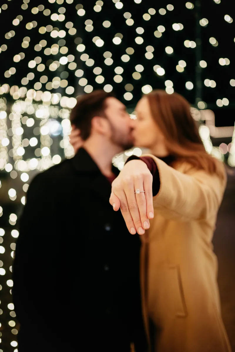 A newly engaged couple kissing beneath strings of lights, the ring held toward the camera