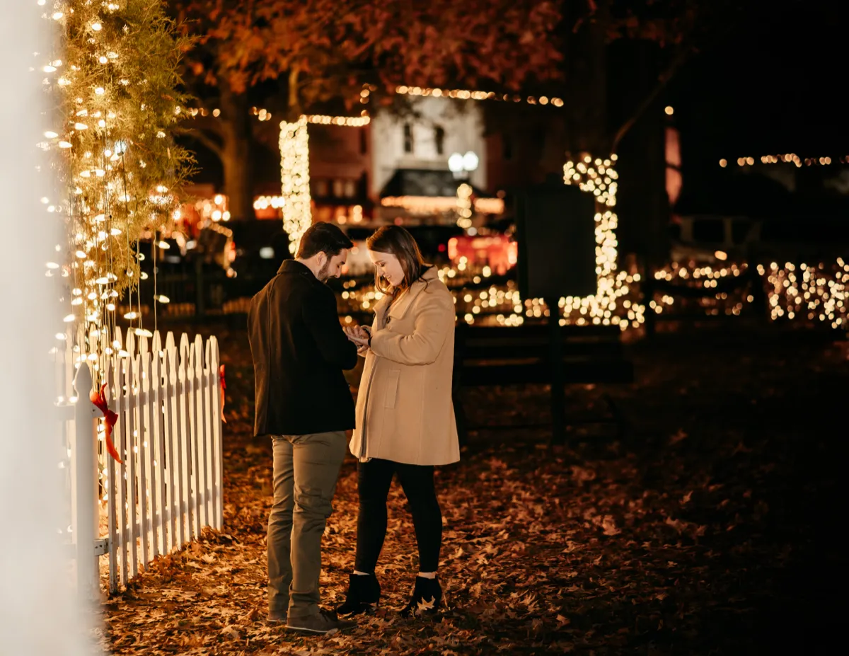 A couple looking at the ring together on a lawn strung with holiday lights