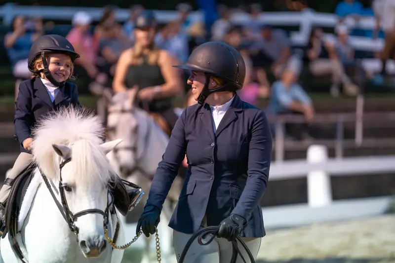 A small child on a white pony sharing a smile with an adult rider beside her at a show