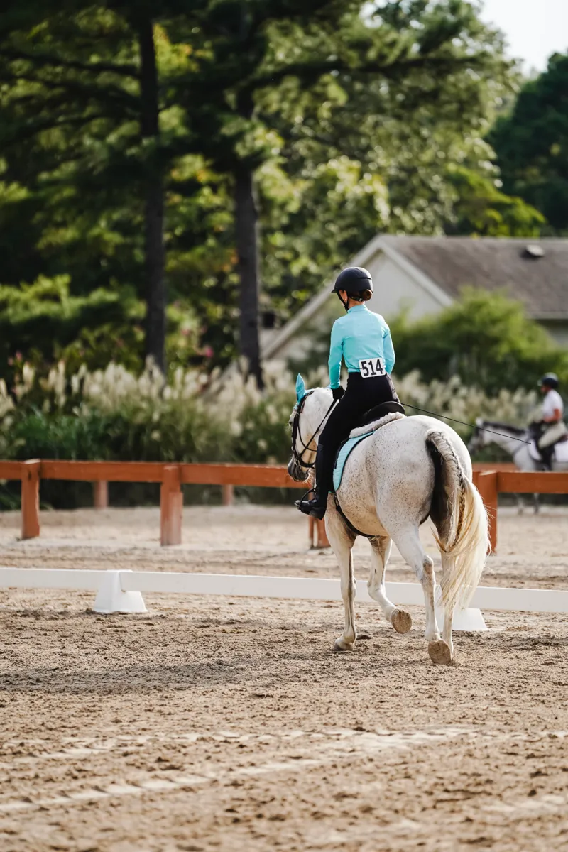 A rider in teal riding a grey horse away down a dressage arena
