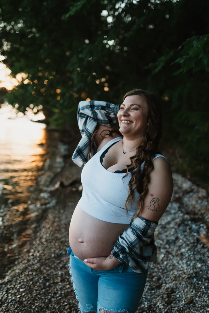 An expectant mother in a flannel shirt laughing at the shoreline at golden hour