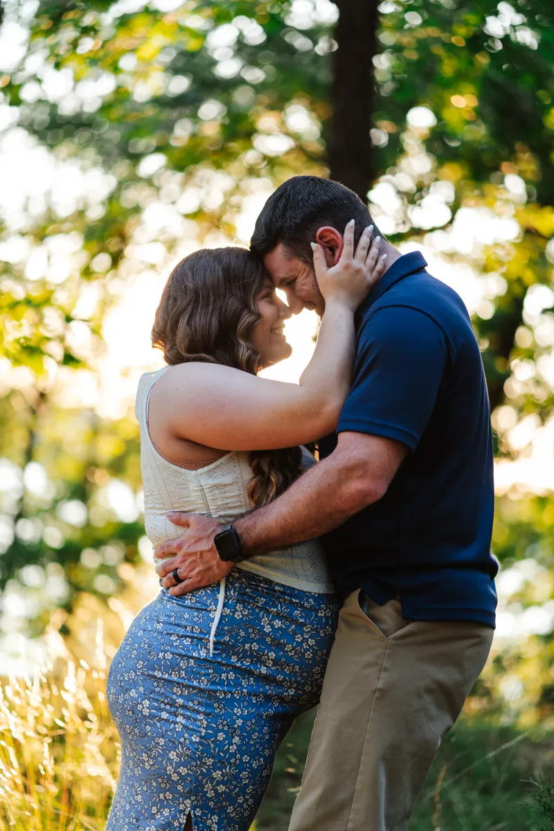 An expectant couple standing forehead to forehead in tall grass, backlit by evening sun