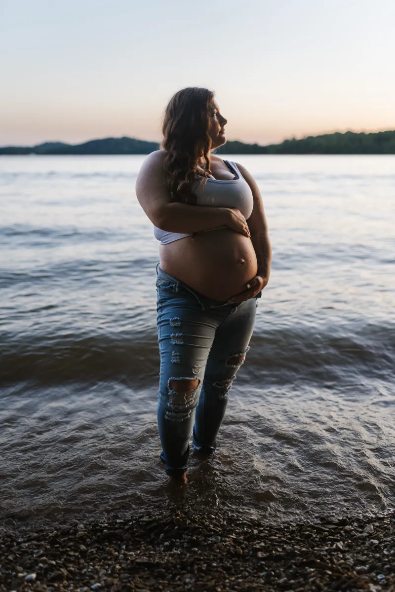 An expectant mother standing barefoot in shallow lake water at dusk
