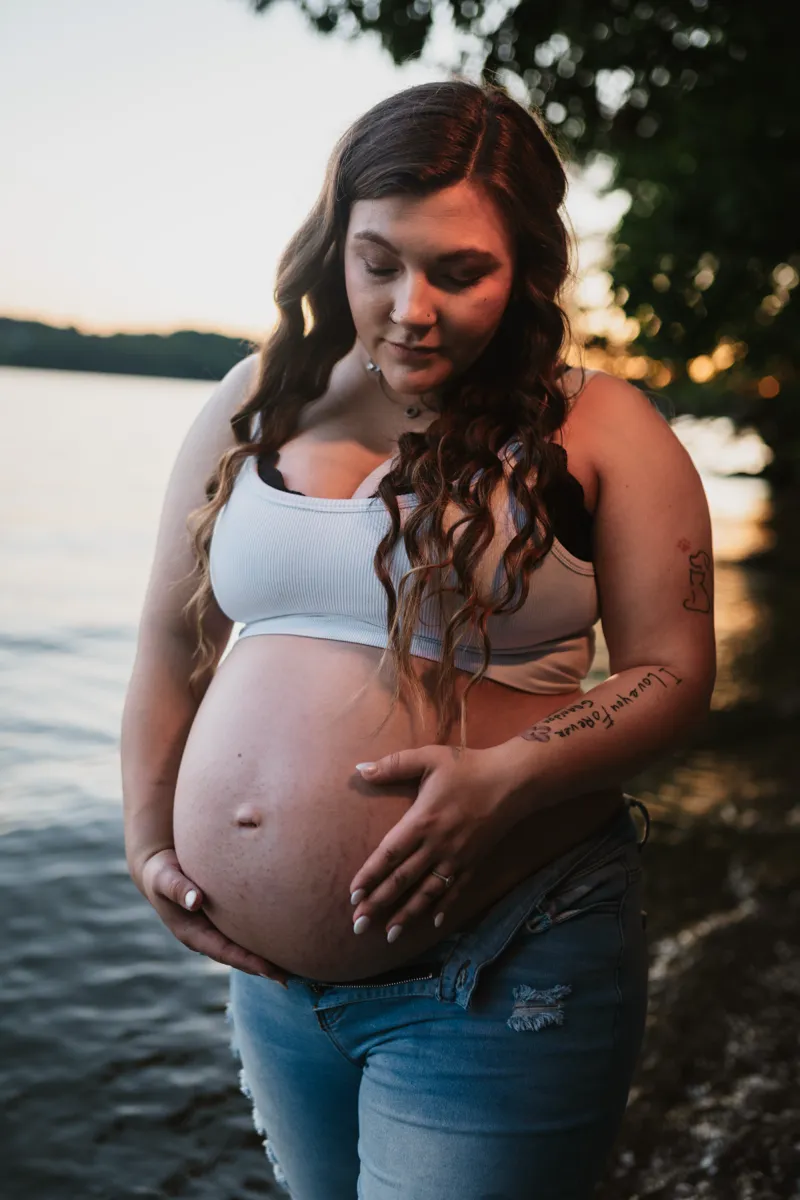 An expectant mother looking down at her belly, lake and evening light behind her