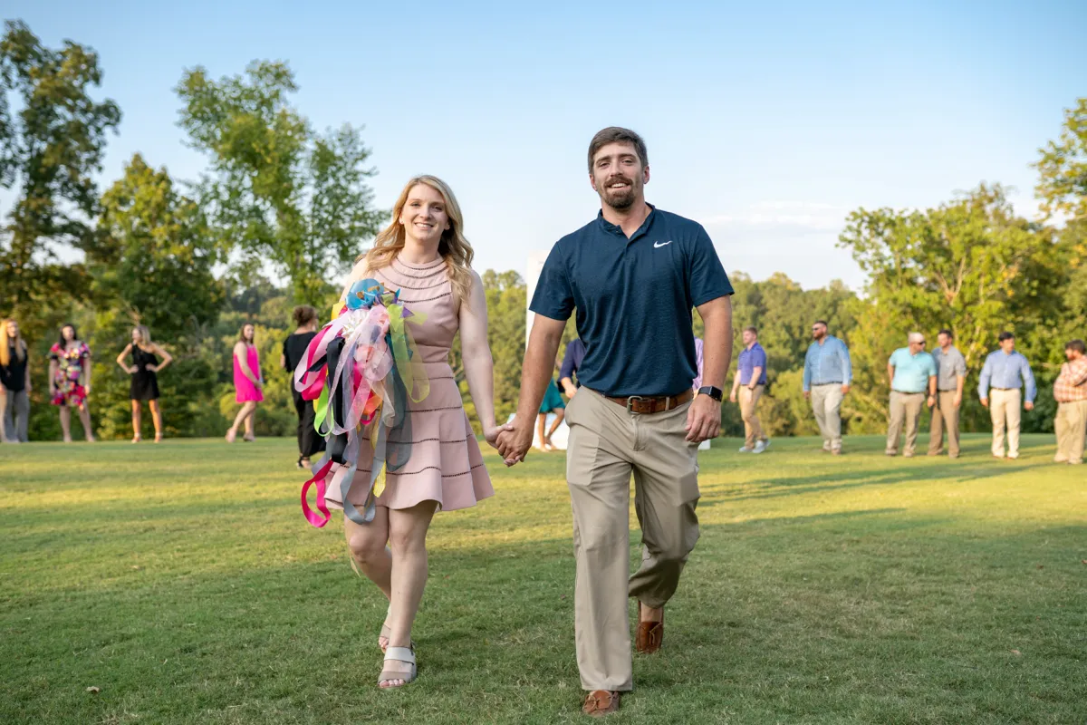 A couple walking hand in hand across a lawn as guests look on