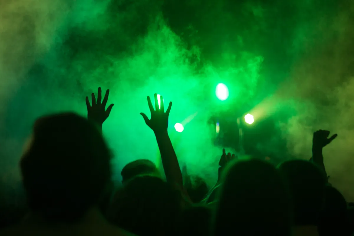 Hands raised in silhouette against green stage light and haze on a dance floor