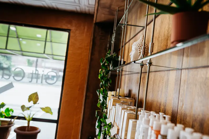 Glass shelves of white skincare products and a ceramic Buddha head against a warm wood wall, beside a bright front window and potted plants