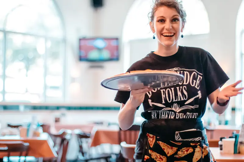 A smiling server holding out a pizza on a metal tray in a bright restaurant dining room with arched windows