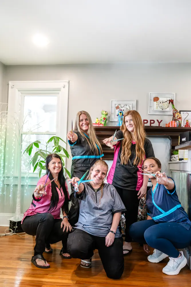 Five groomers crouched together on a salon floor, holding their shears out toward the camera and laughing