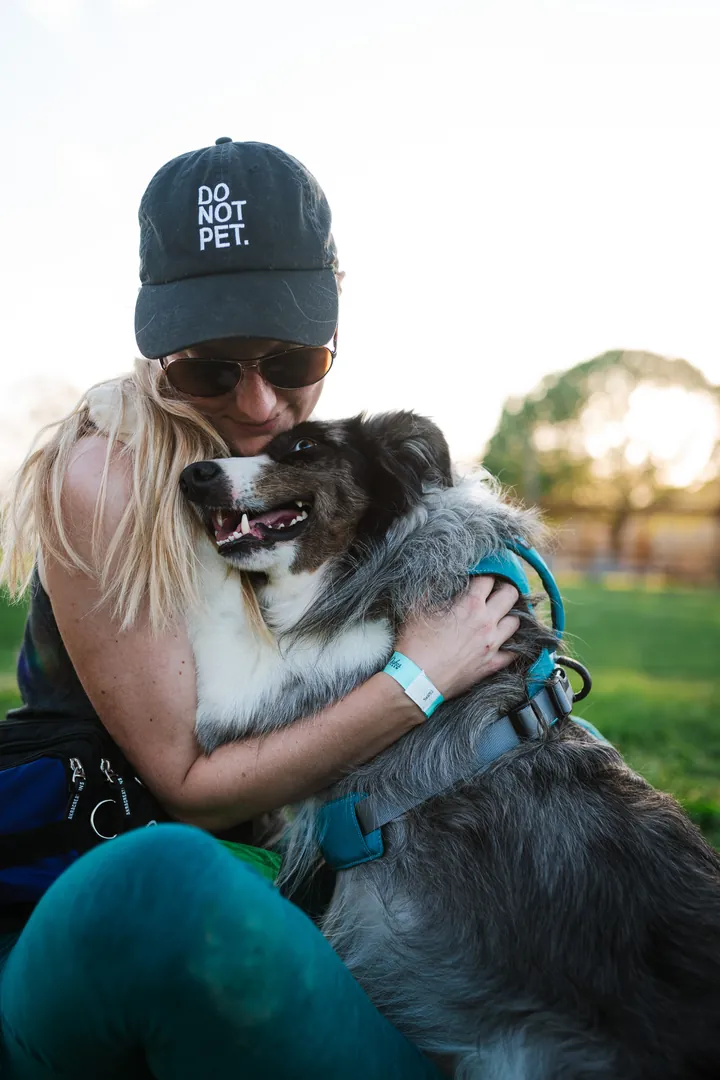 Sara Carson in a DO NOT PET cap and sunglasses hugging a grey merle dog close on the grass at golden hour