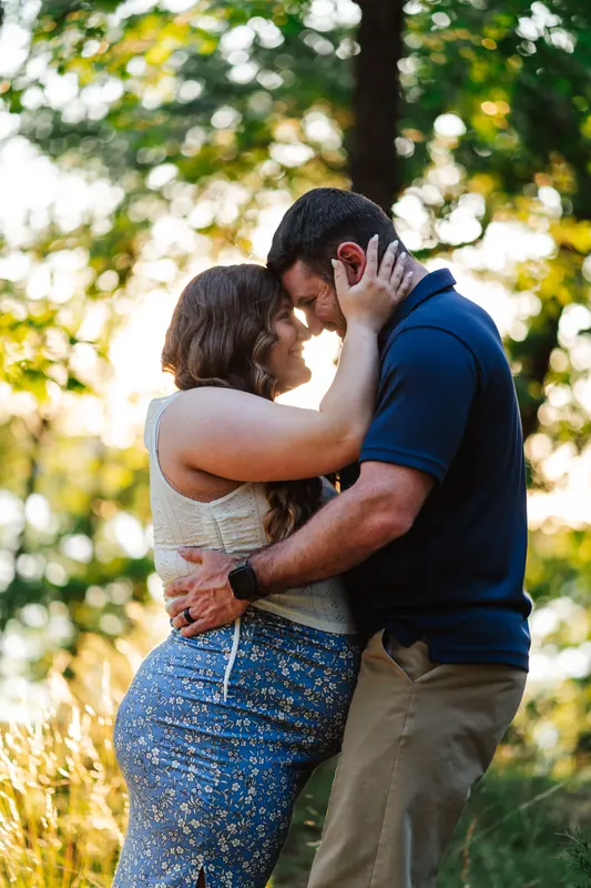 A couple embracing forehead to forehead under the trees in golden evening light, his hand resting on her cheek
