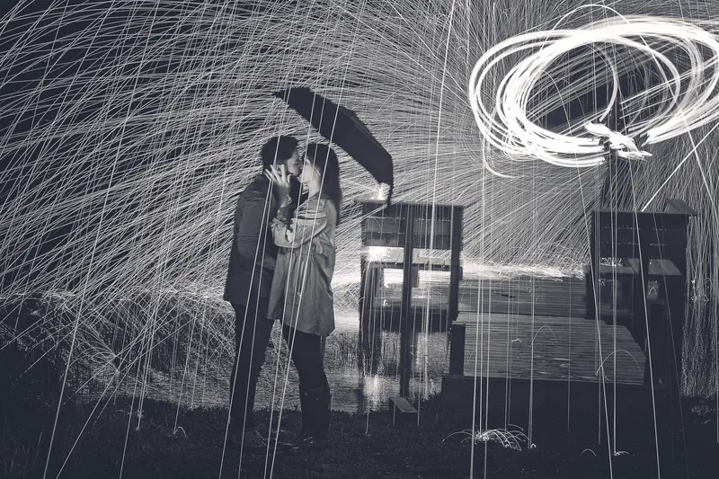A black-and-white night portrait of a couple kissing under an umbrella beside a lake dock, showered in arcs of steel wool sparks